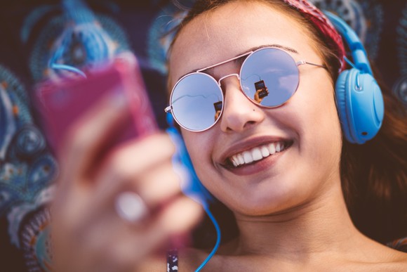 A smiling person wearing sunglasses listens to music on a phone.
