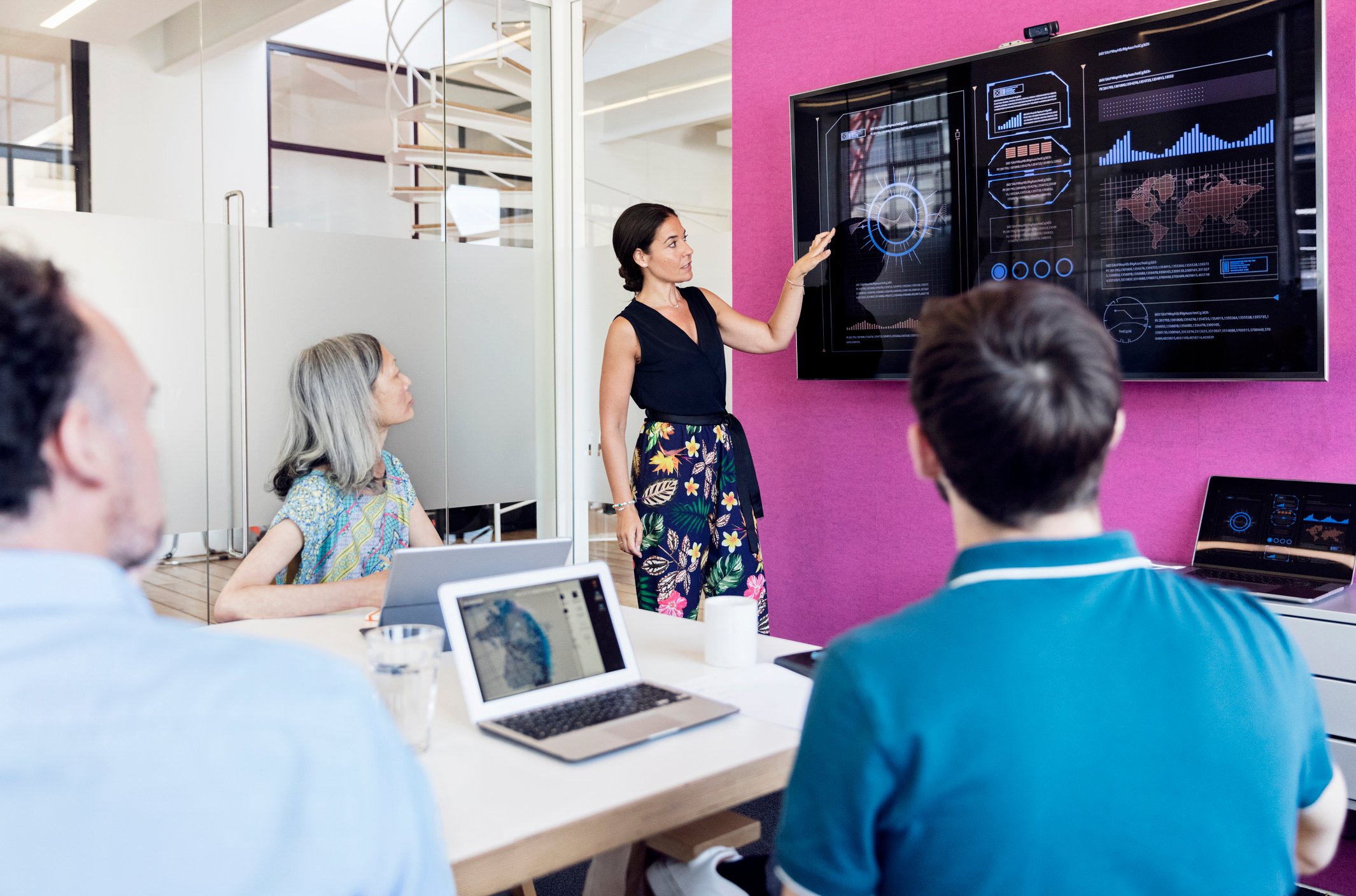 A meeting room with people looking at a large screen of data and graphs on the wall.