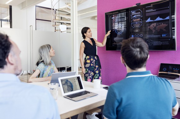 A meeting room with people looking at a large screen of data and graphs on the wall.