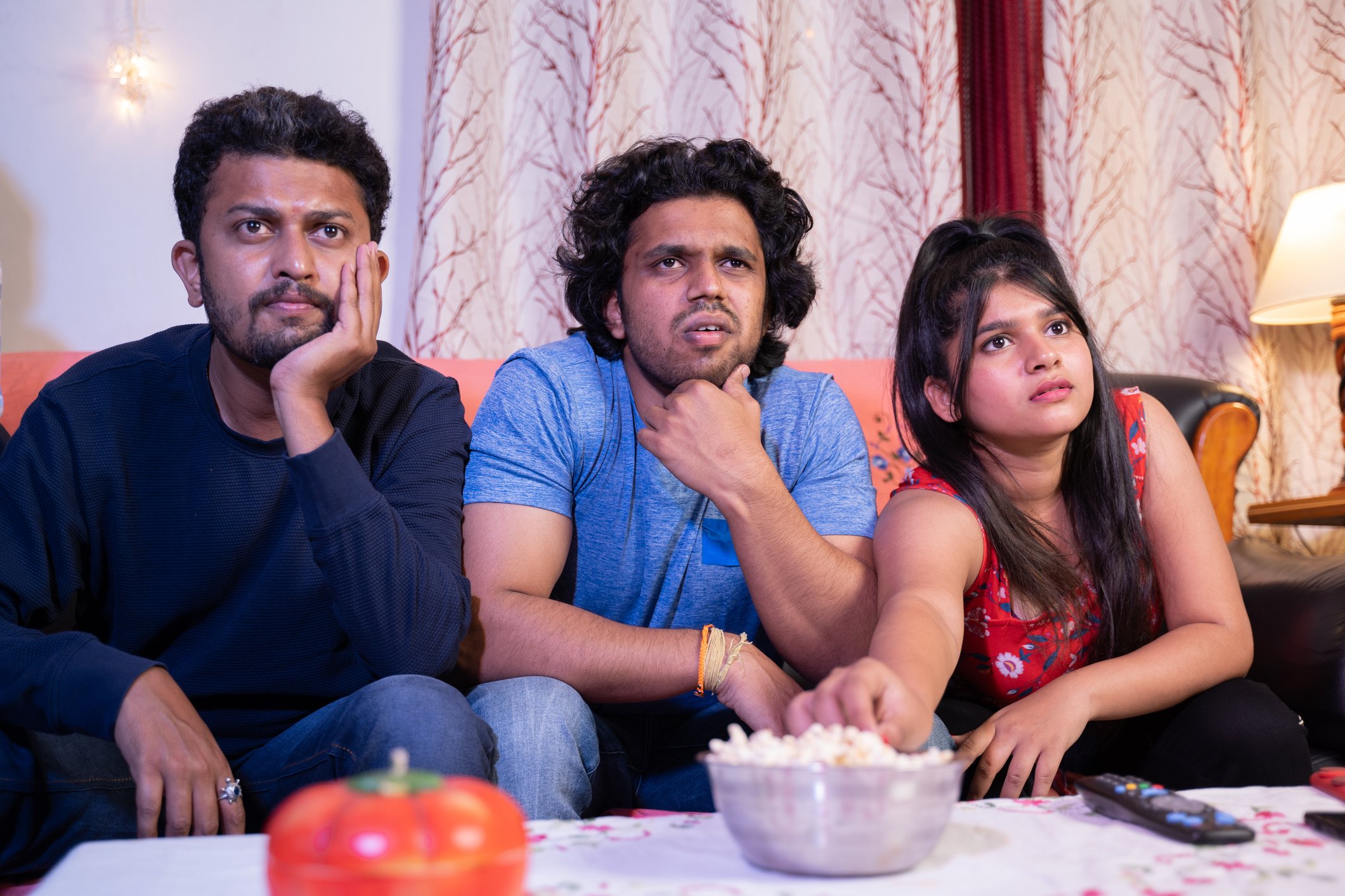 Three people sitting on couch, eating popcorn, and watching TV.