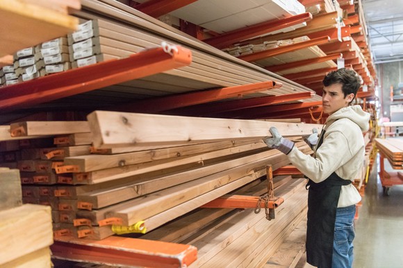 A man wearing gloves, a hoodie, and an apron lifts a piece of lumber off a stack of boards in a warehouse full of cut lumber supplies