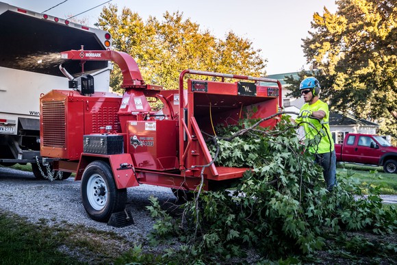 A chipper, made by Alamo's subsidiary Morbark, being used by a man in a blue hard hat and yellow warning vest to chip branches.