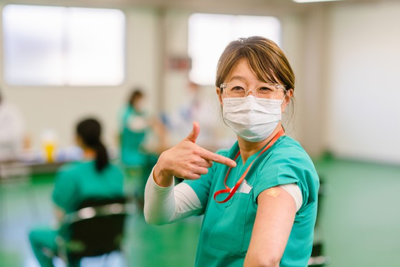A healthcare worker points to a bandage on their upper arm indicating a vaccination.