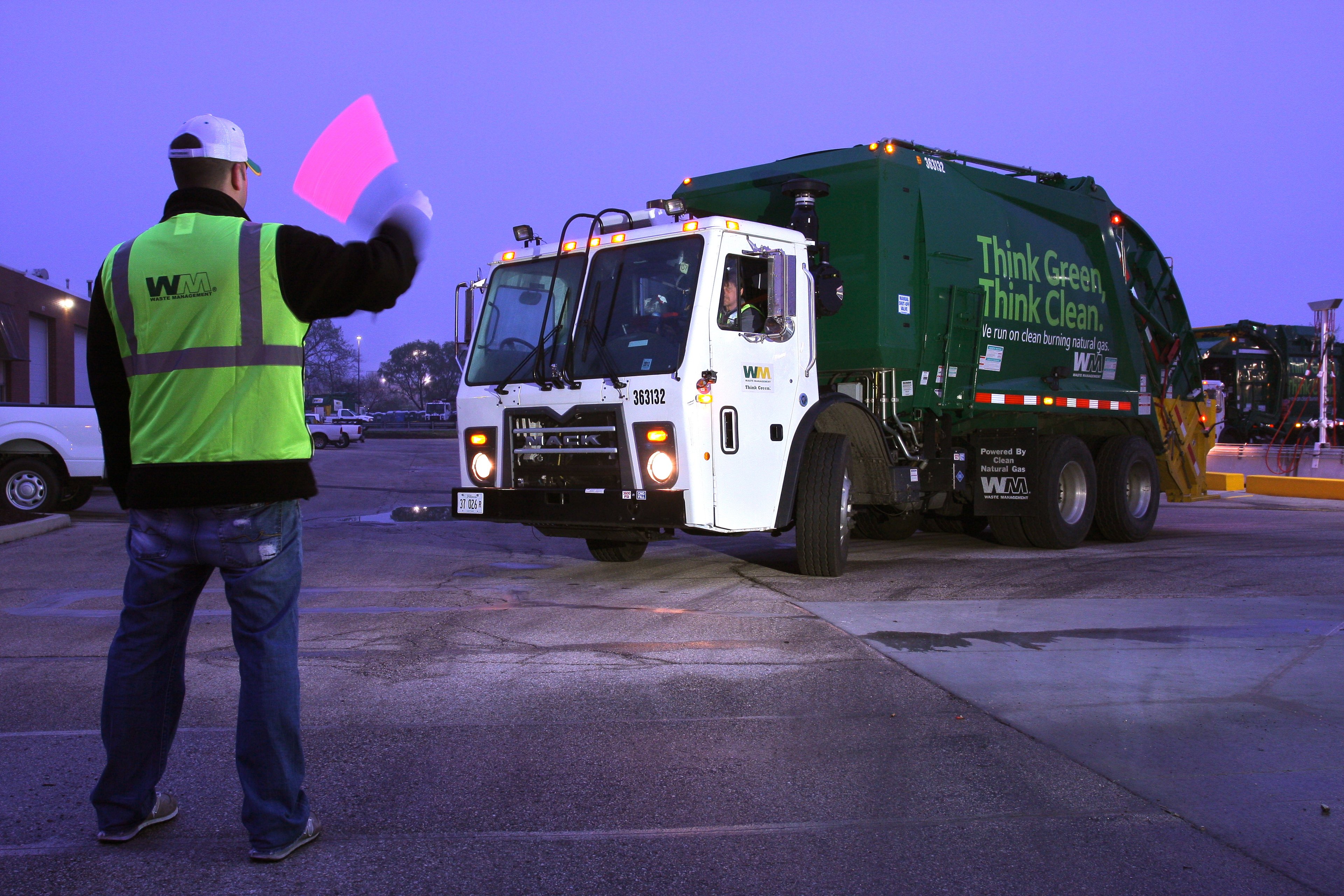 A man with a flag directs a garbage truck as it leaves its lot at dawn.