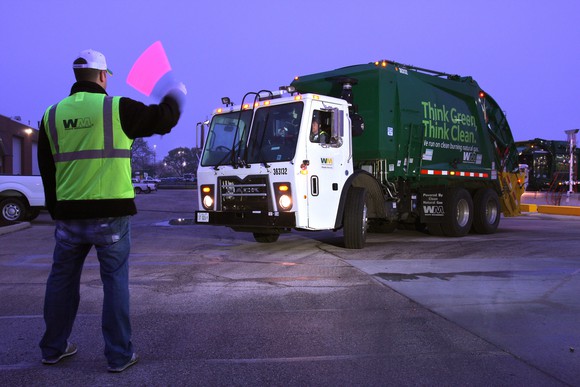 A man with a flag directs a garbage truck as it leaves its lot at dawn.
