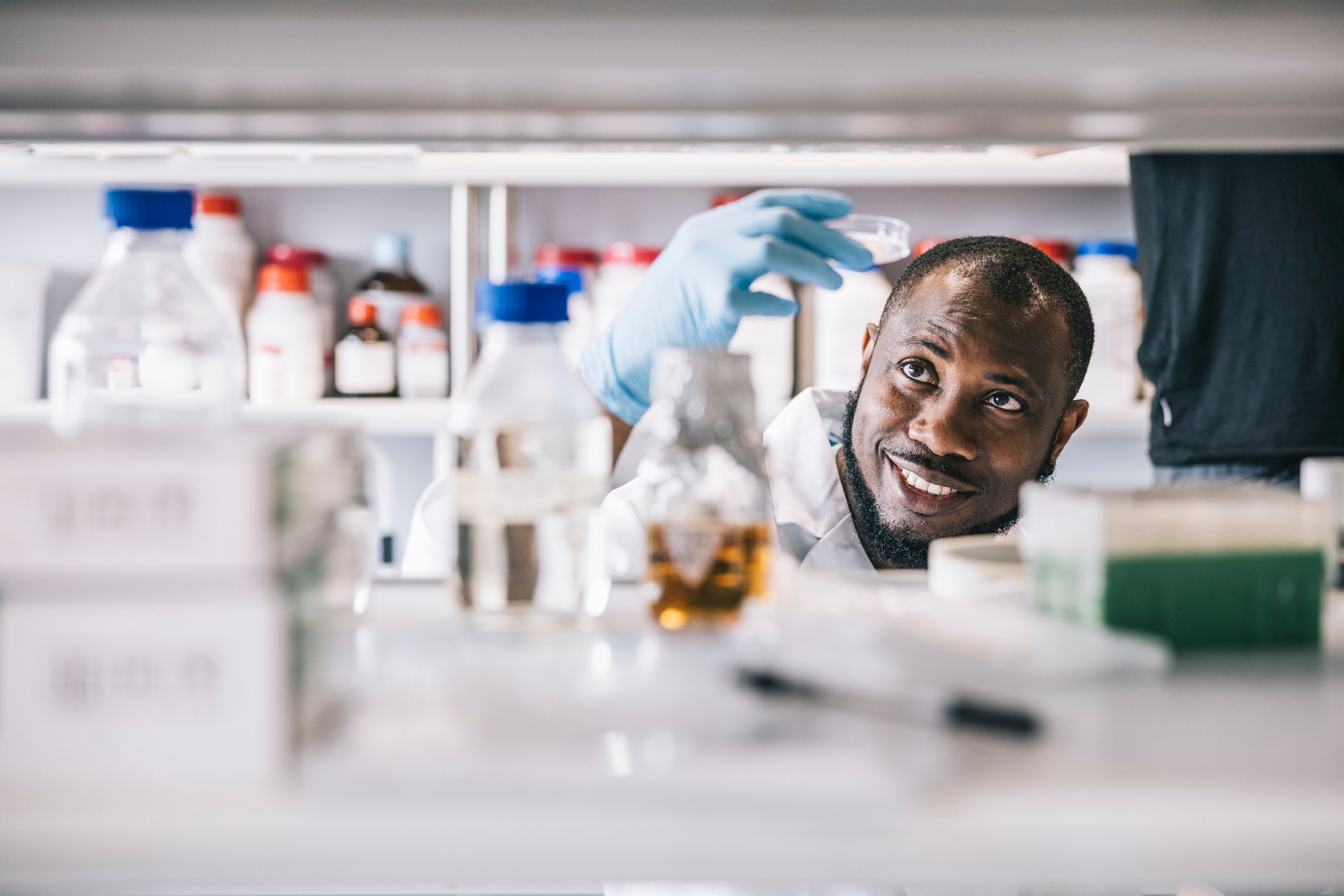 A smiling scientist holds up a petri dish to the light while working at a cluttered laboratory bench.