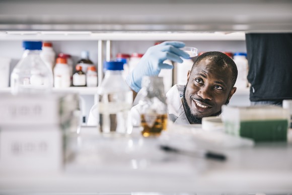 A smiling scientist holds up a petri dish to the light while working at a cluttered laboratory bench.
