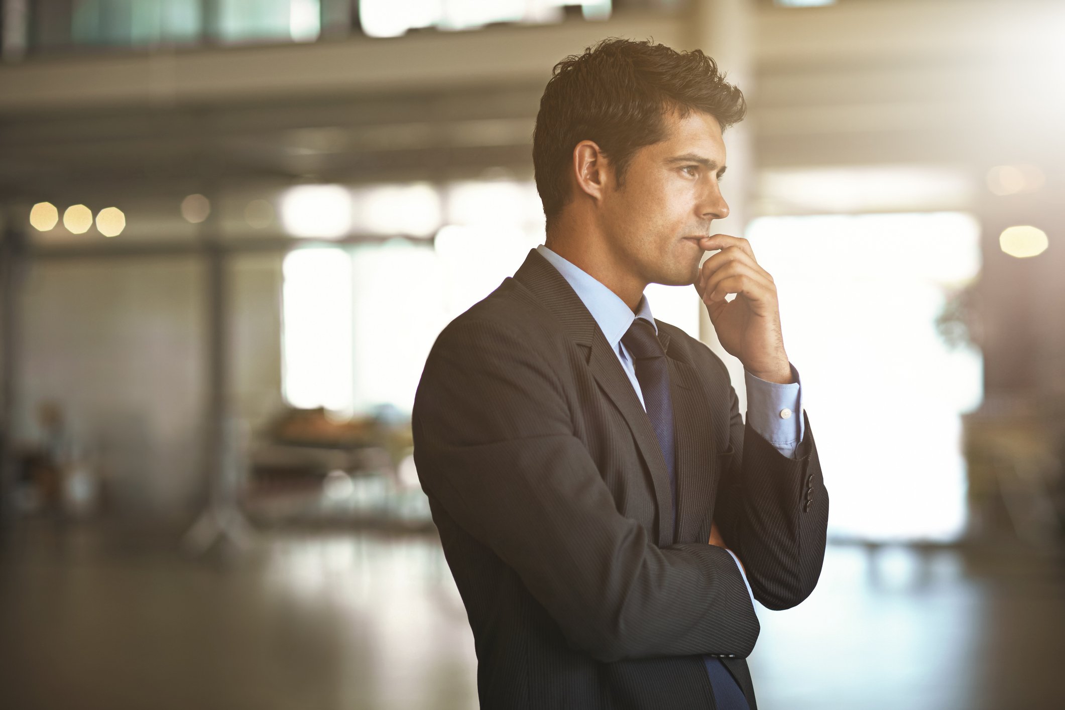 A person in a business suit looks concerned while standing in a lobby and looking off into the distance.
