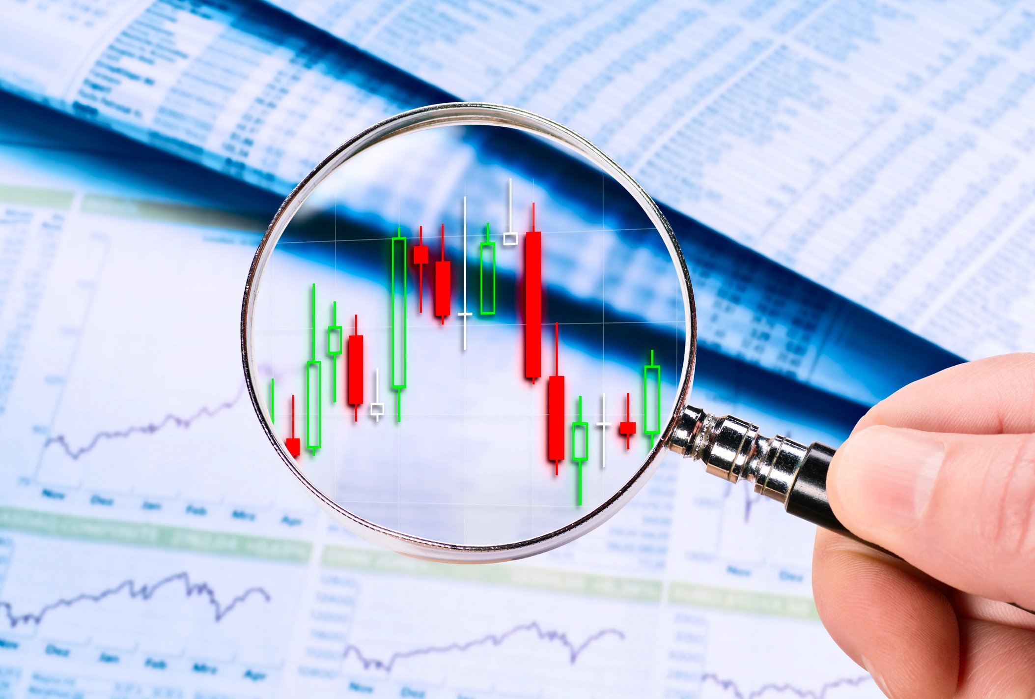 A hand holds a magnifying glass over a volatile stock chart.