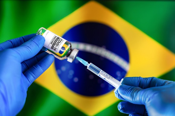 Gloved hands holding a syringe and a COVID-19 vaccine vial in front of a Brazilian flag.