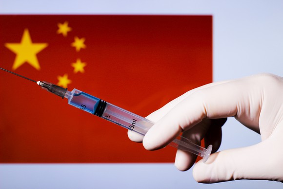 Gloved hand holding a syringe with needle with a Chinese flag in the background.