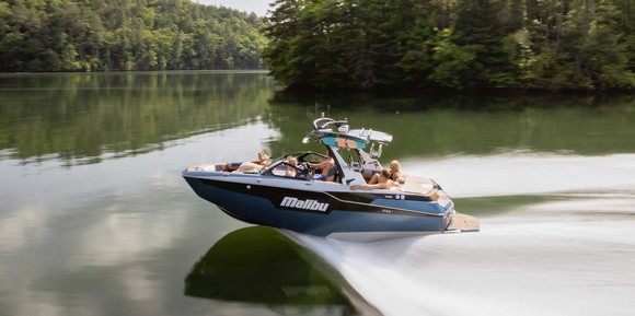 A Malibu M220 ski boat races across the water of a lake with trees being seen on the shoreline. The boat has several passengers in it