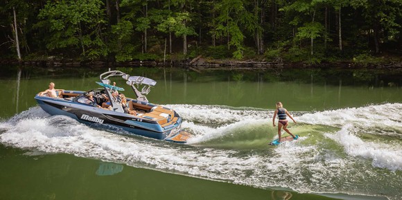 A Malibu M220 boat motors in a waterway with passengers aboard and a water skier being towed behind it