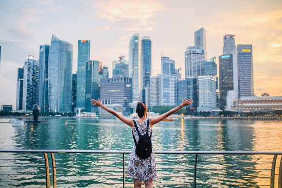 Young person wearing a backpack, arms outstretched, facing the skyline across Singapore Bay.