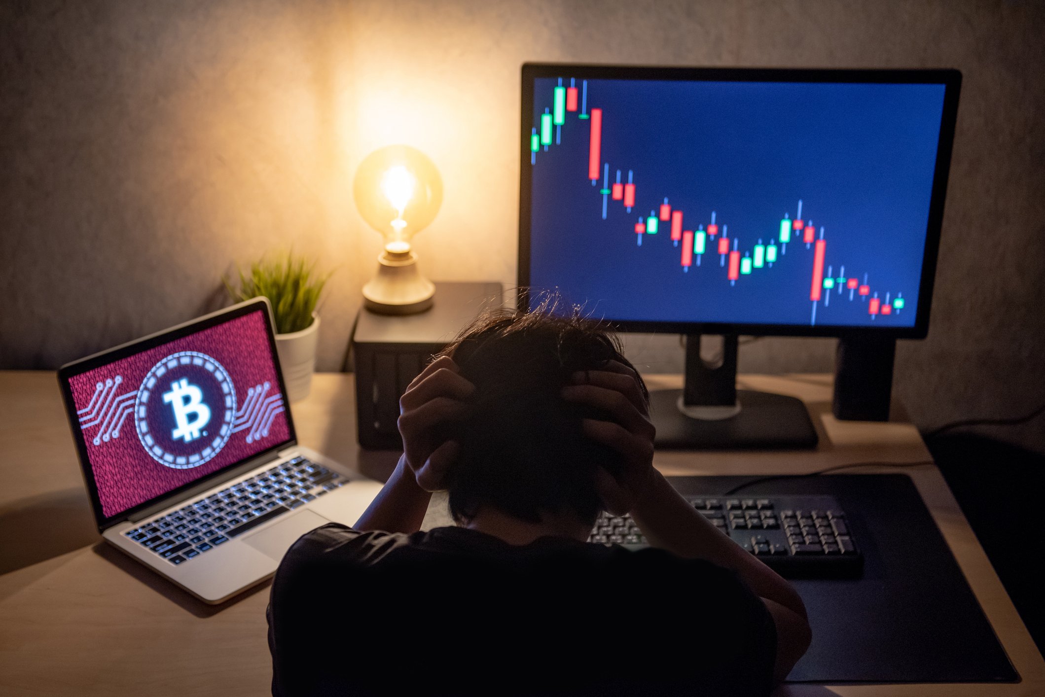 Trader in distress at desk with declining stock chart on one screen and bitcoin logo on the other.