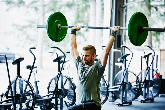 Someone doing barbell lunges at a gym with exercise machines lined up behind them.