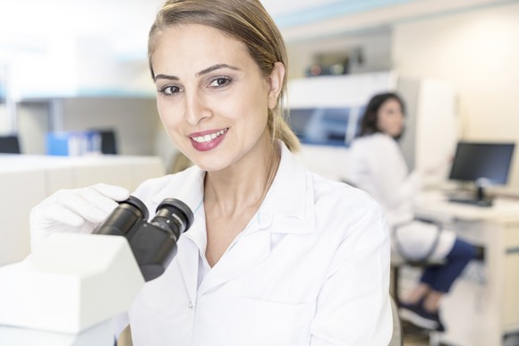 A researcher in a lab in front of a microscope looking into the camera.