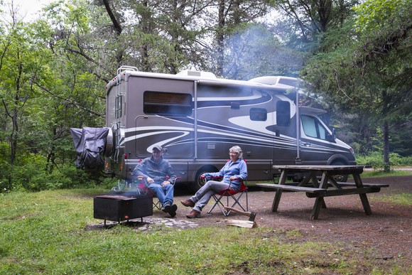 Two people sitting outside their RV camper in a forested campground with a fire.