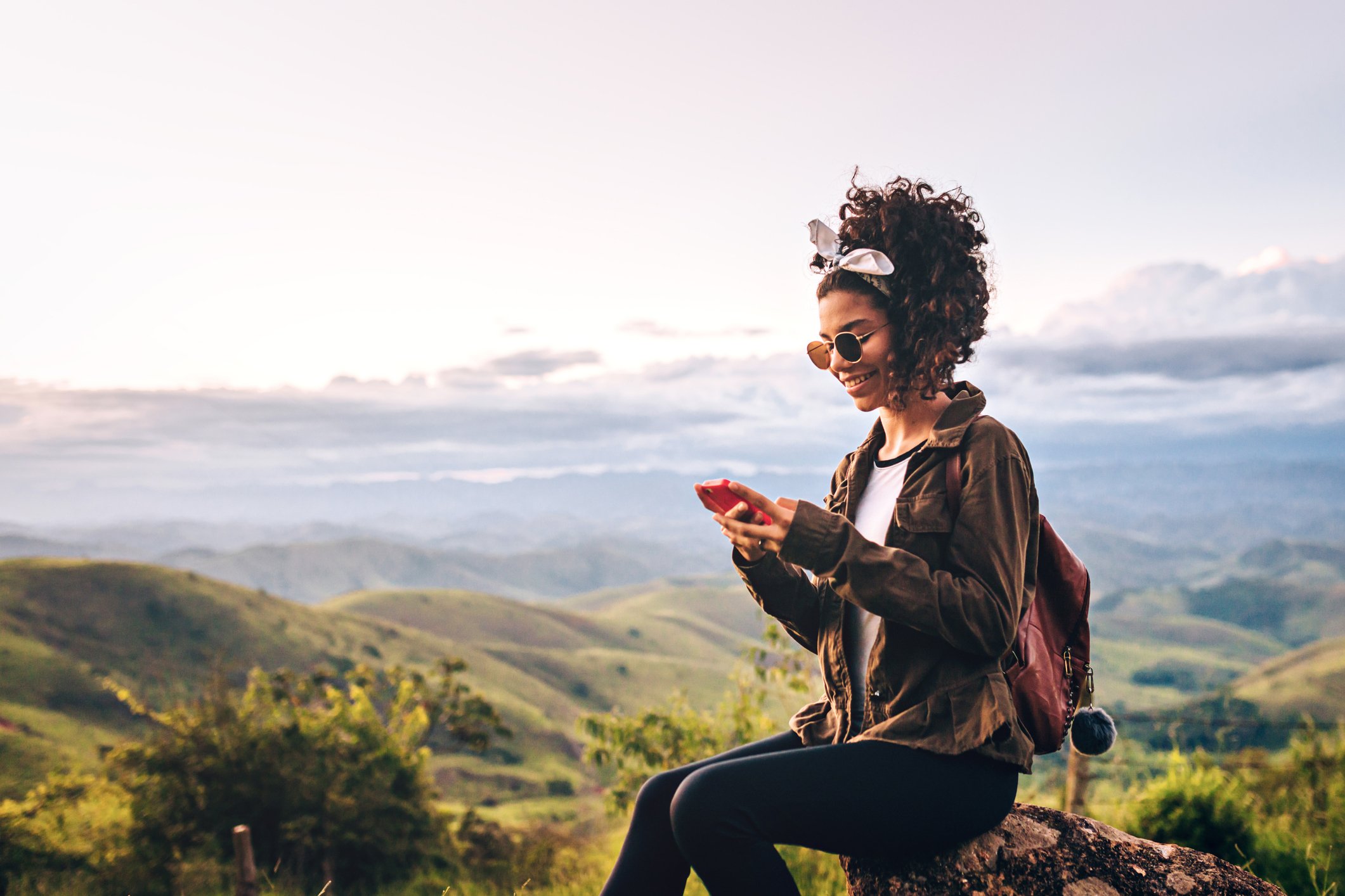 Hiker sits on a rock and accesses phone service from a mountaintop.