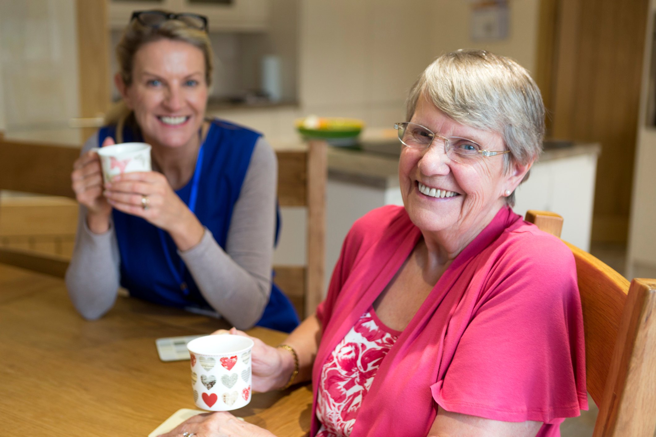A healthcare worker joins an an elderly woman for a cup of tea while sitting at a kitchen table.