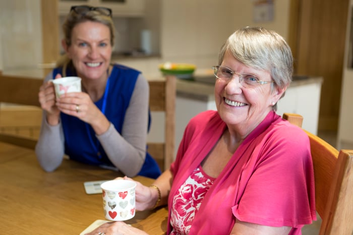 A healthcare worker joins an an elderly woman for a cup of tea while sitting at a kitchen table.