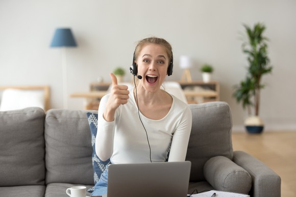 A person wearing an audio headset at her laptop gives a thumbs-up to the camera.