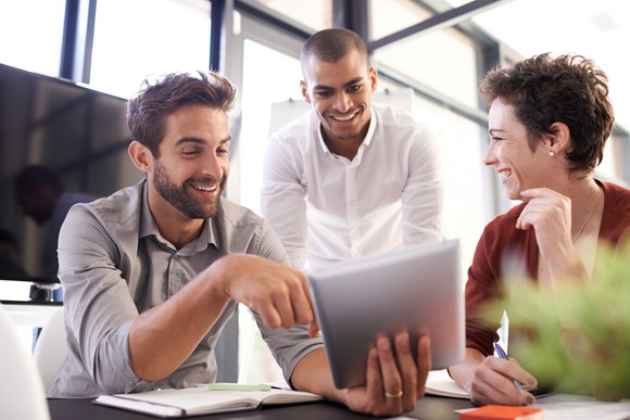 Three colleagues smiling and working on a tablet in an office.