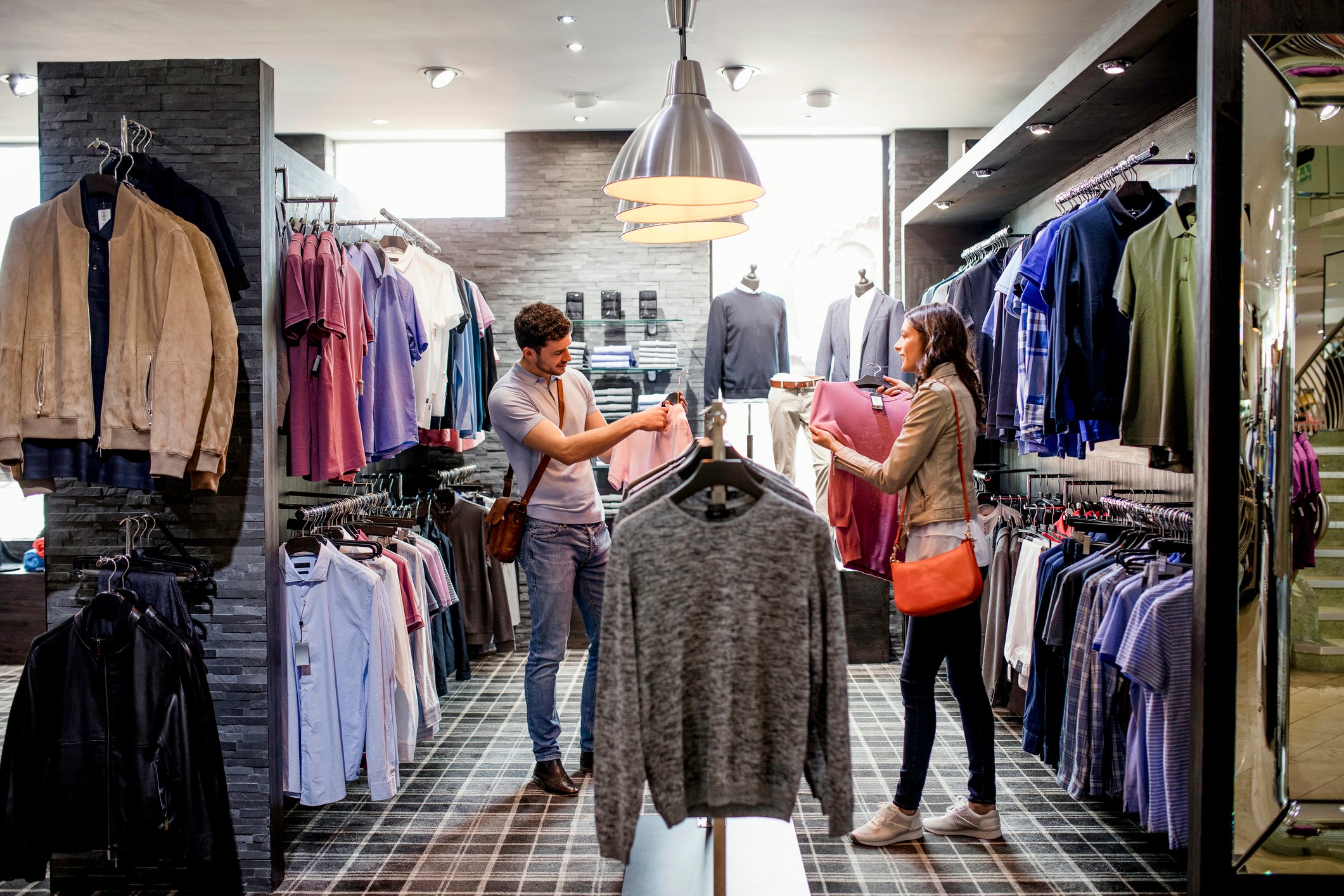 Two people shopping for clothing in a store, surrounded by racks of tops and jackets.