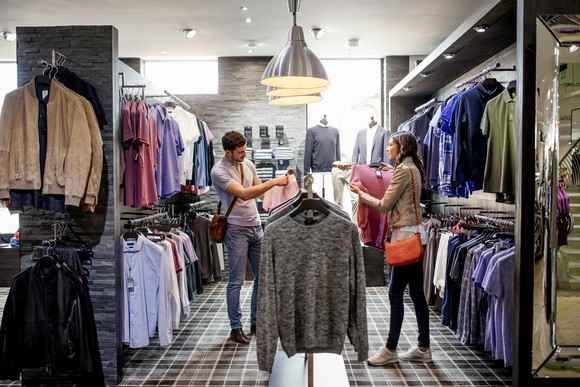 Two people shopping for clothing in a store, surrounded by racks of tops and jackets.