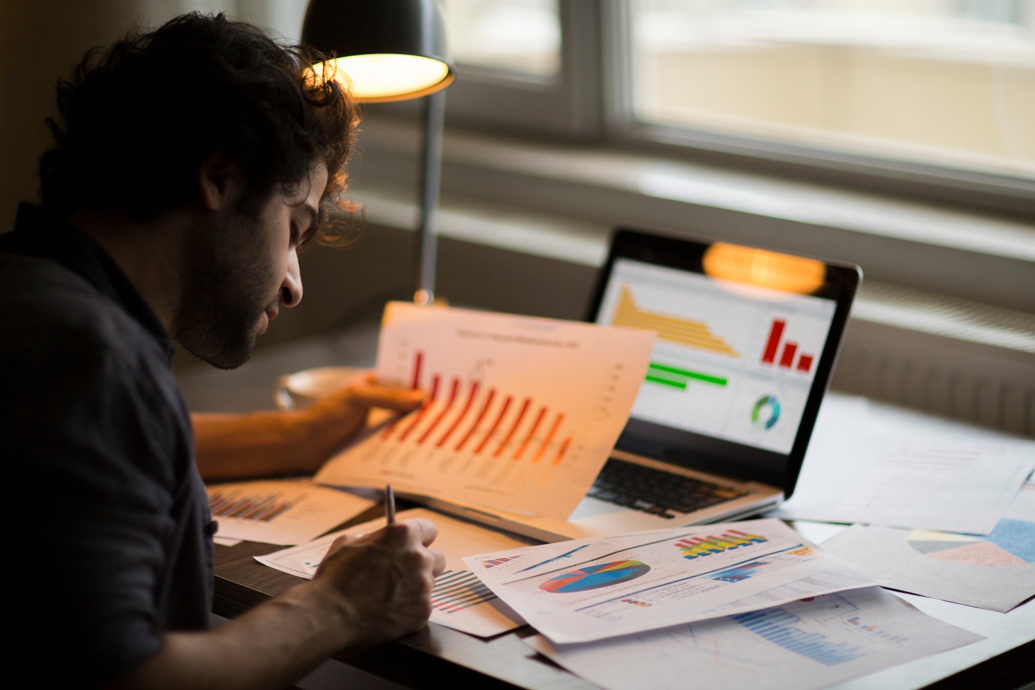 A man sits at a desk under lamp light looking at charts and graphs on pieces of paper with a laptop computer sitting in the background