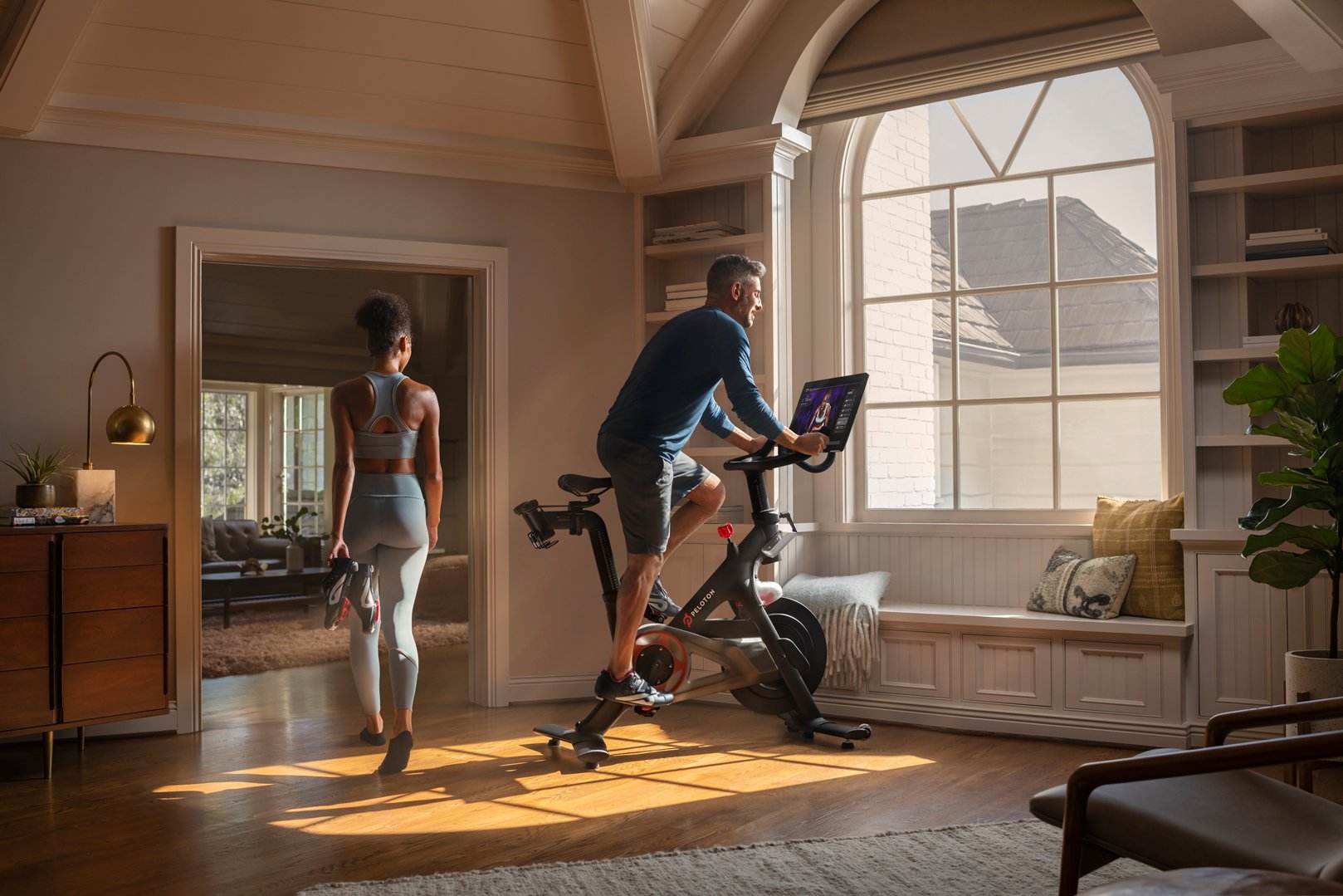 A man exercises on a Peloton bike in his home while looking out a large window as a woman wearing exercise clothing walks by inside the house.