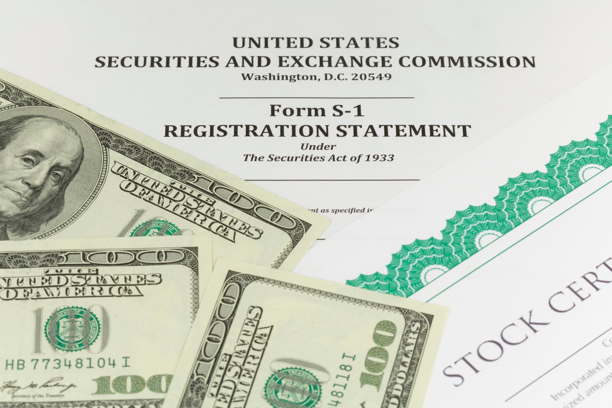 A paper S-1 registration statement form for an IPO, with a stock certificate and several hundred-dollar bills lying on top of it.  