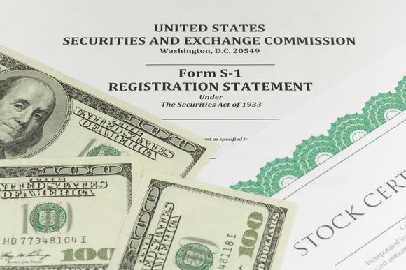 A paper S-1 registration statement form for an IPO, with a stock certificate and several hundred-dollar bills lying on top of it.  