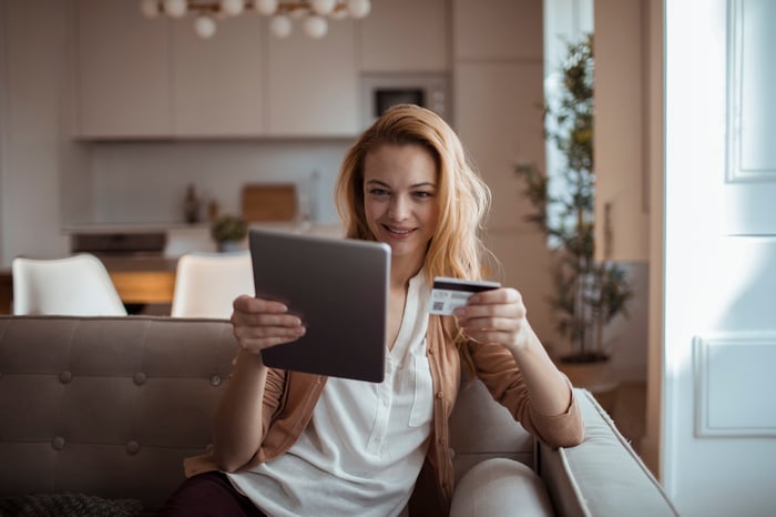 Young woman holds up a tablet and credit card while reclining on a couch. 