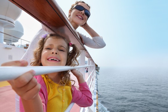 Adult and child smile and lean against the railing of a cruise ship at sea.