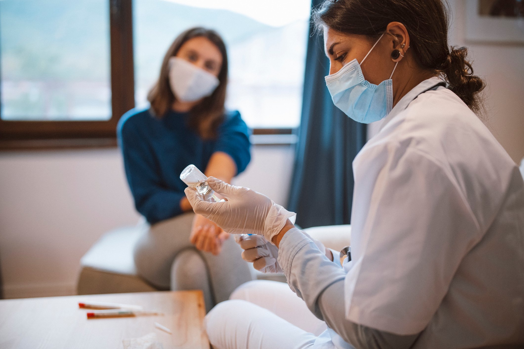 A healthcare worker draws up a vaccine while a patient waits in the background.