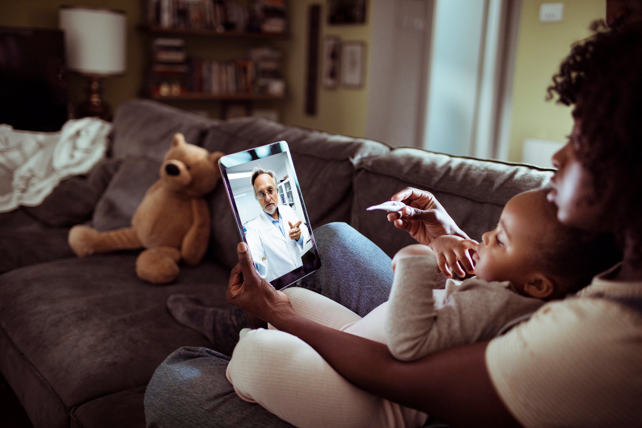 An adult holds an infant while participating in a virtual visit with a physician.