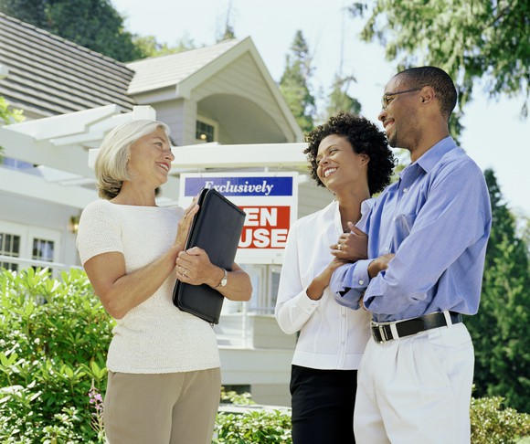 A real estate broker and a couple stand in front of an "Open House". sign.