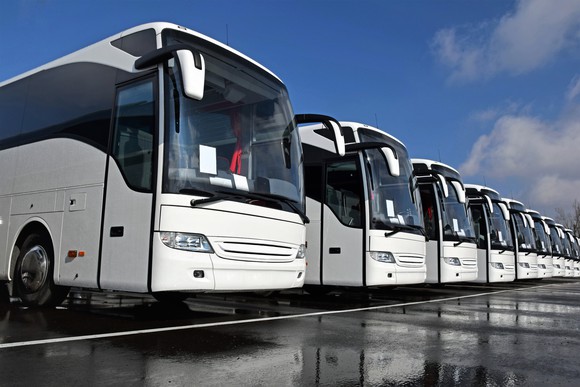 A row of white tour buses lined up outside a factory on wet pavement after being manufactured.