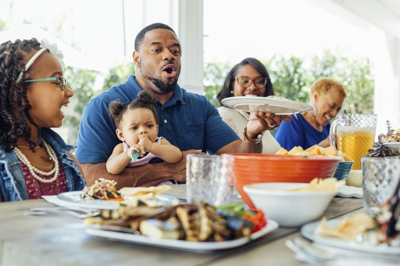 A group of adults and kids seated at a table and partaking of barbecue food.