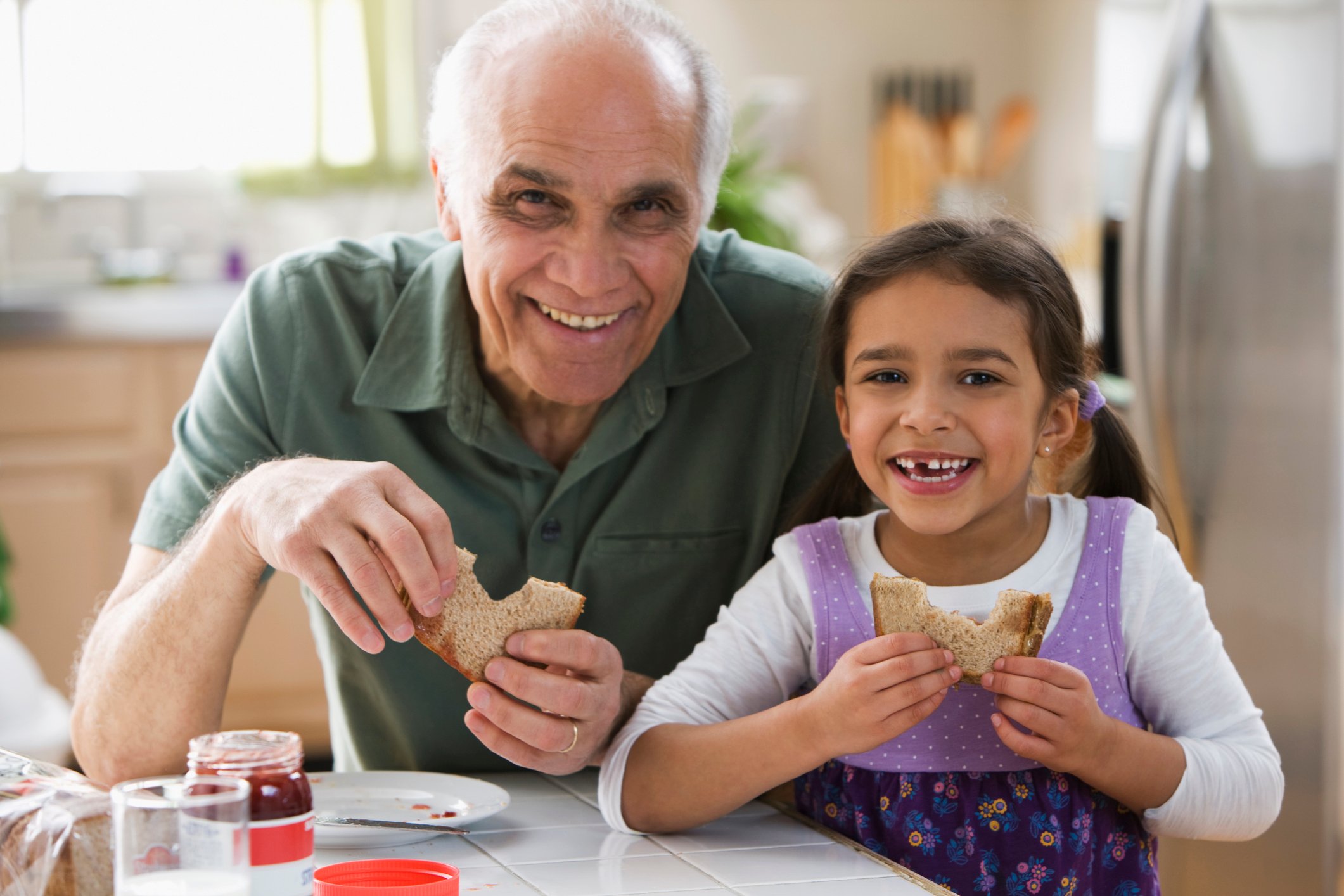 A smiling adult and child enjoying peanut butter and jelly sandwiches.