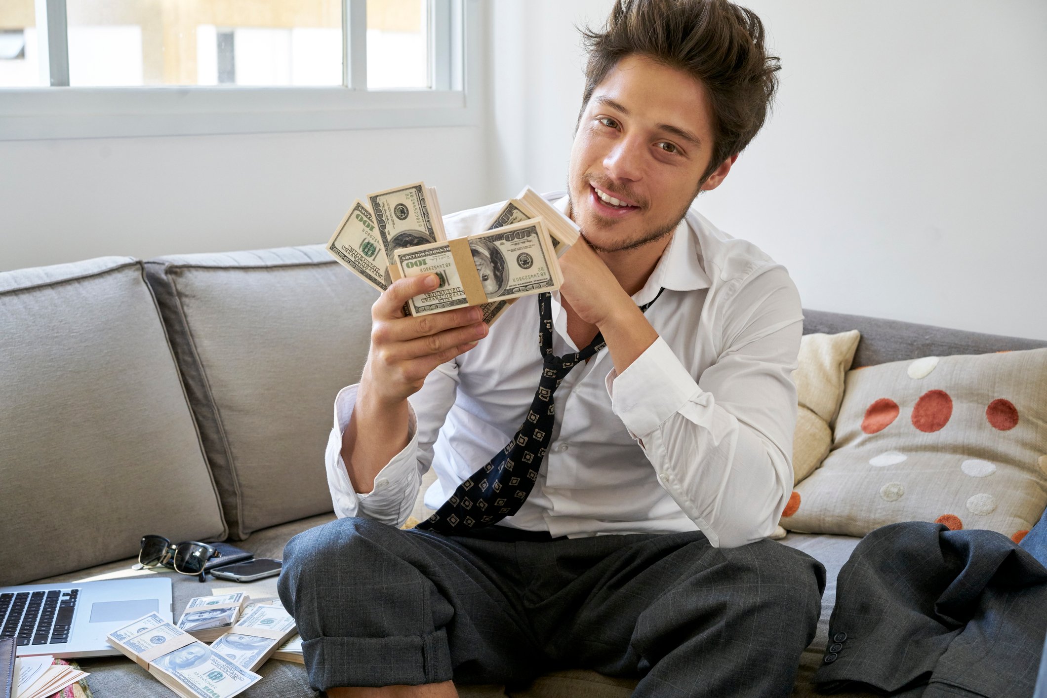 A disheveled man sits on a sofa holdings stacks of cash.
