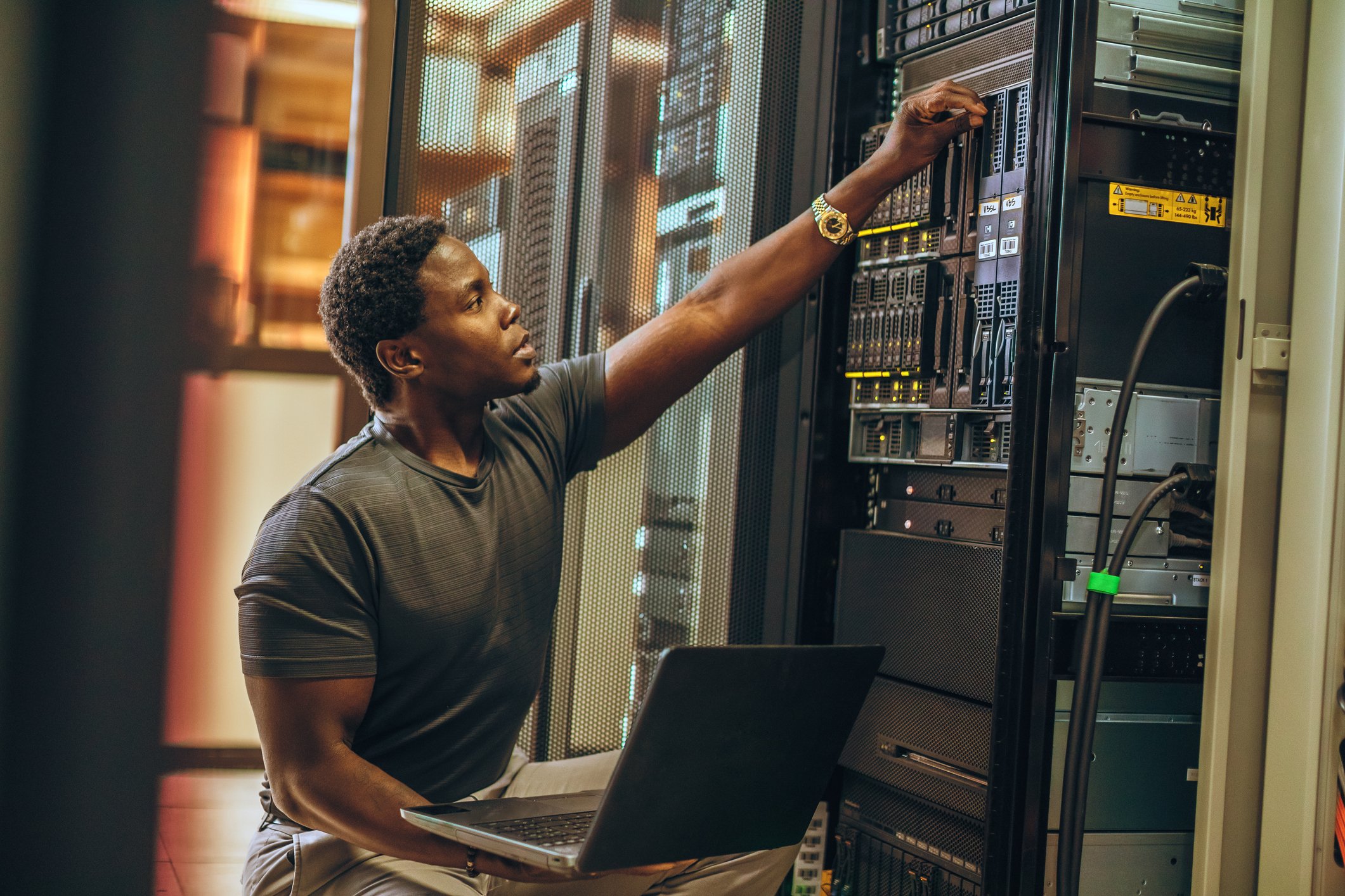 young technician in server room has laptop on his lap. 