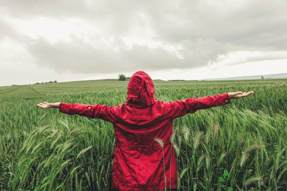 A person in a red jacket standing in a field with outstretched arms as a storm rolls in.