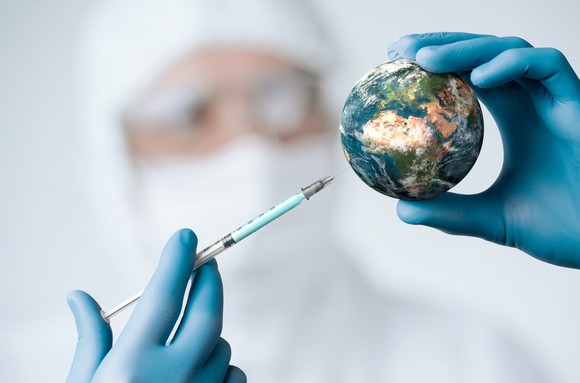 A researcher holds up a syringe to a small globe as if to vaccinate the world.