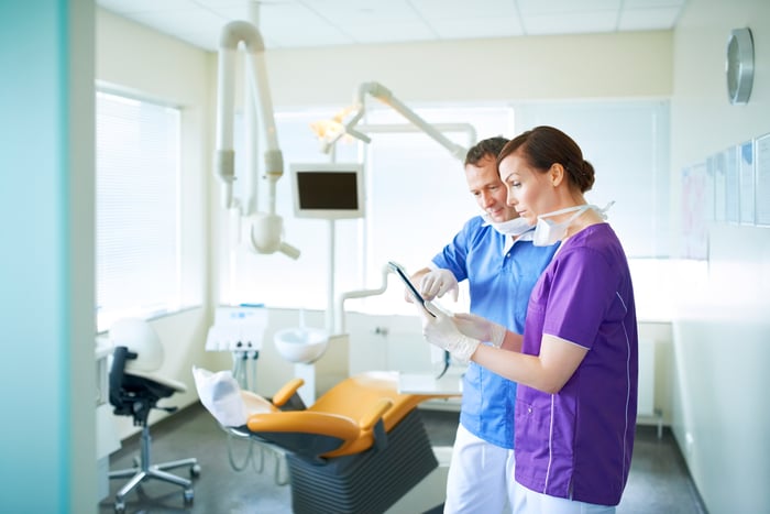 Two dentists examine an image on a tablet while standing in an empty examination room.