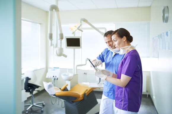 Two dentists examine an image on a tablet while standing in an empty examination room.