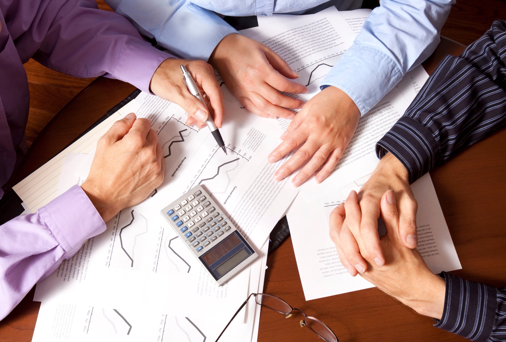 A man wearing a light purple shirt holds a pen that points to a chart sitting on a table along with a calculator as the hands of two older indivduals also rest on the table