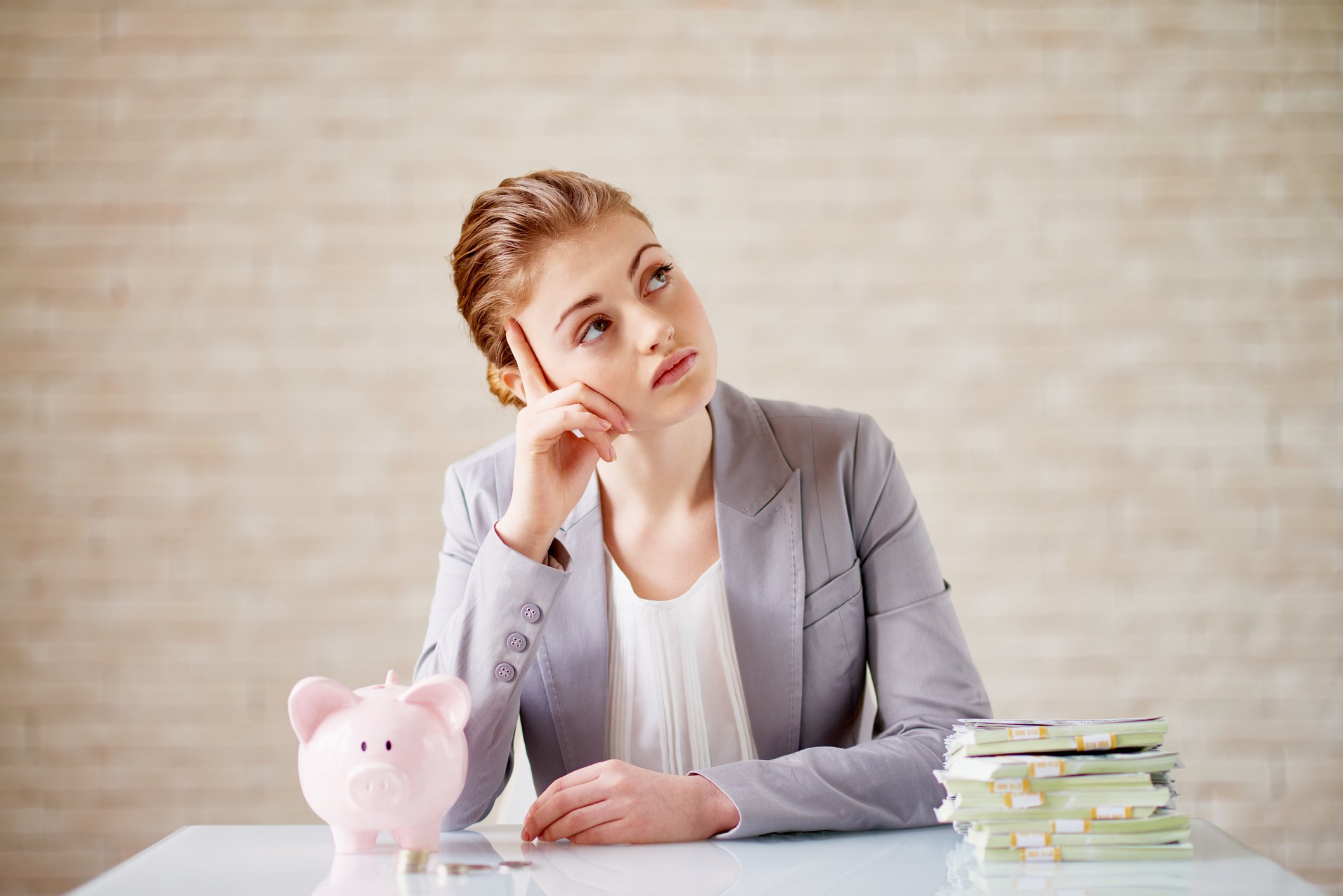A person sits at a table with a piggy bank and a stack of cash and looks upward in thought.