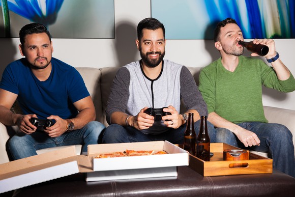 Three people having drinks, eating pizza, and playing video games.
